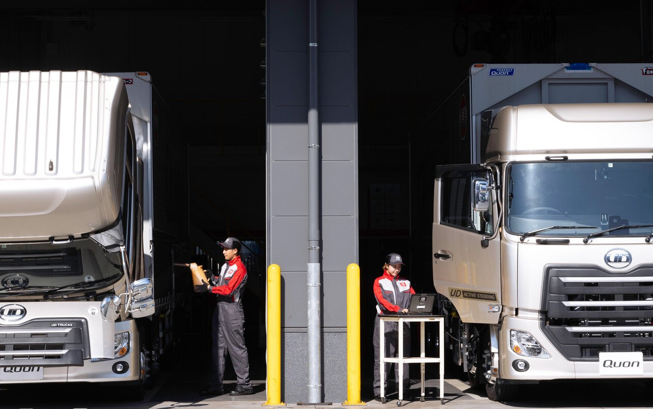 Two workers in red uniforms check two parked delivery trucks in a loading bay.