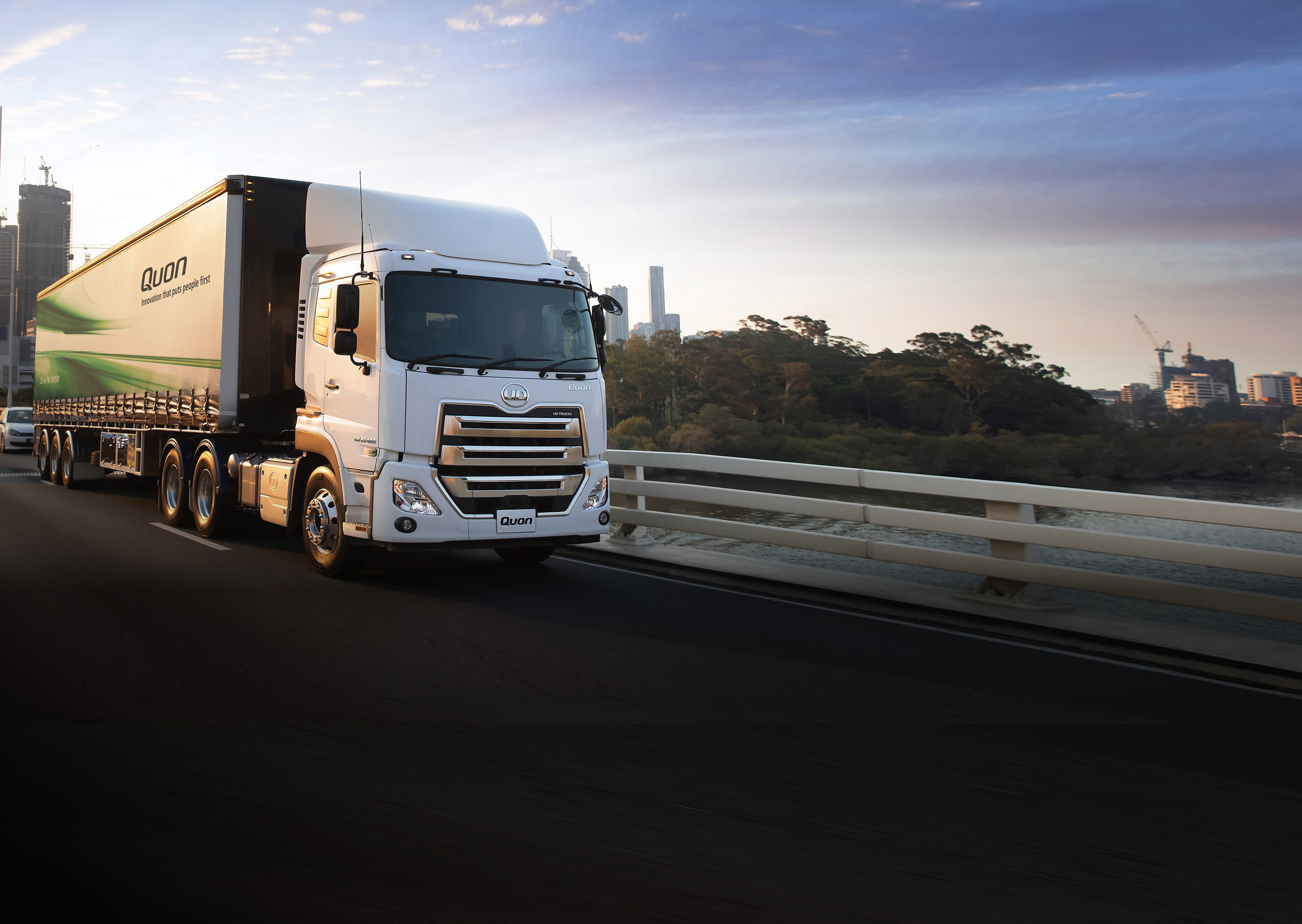 White semi-truck with green accents driving on a city bridge at sunset.