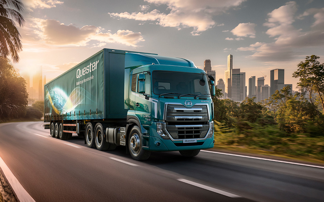 Blue semi-truck driving on a highway with a city skyline in the background at sunset.