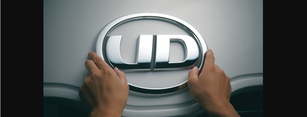 Hands holding a silver UD logo emblem on a gray surface.