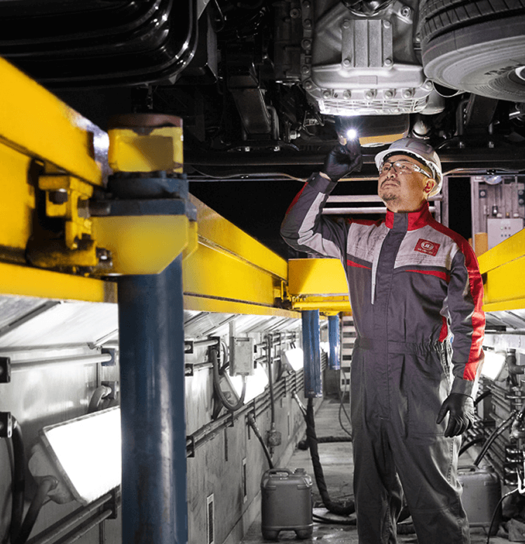 Mechanic in overalls inspecting vehicle underside with flashlight in a garage.