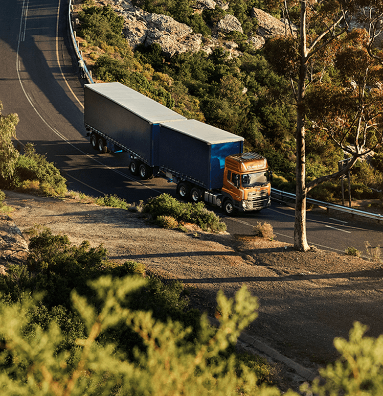 Malaking trak na asul sa kalsadang paliko sa gitna ng mga puno at bundok.