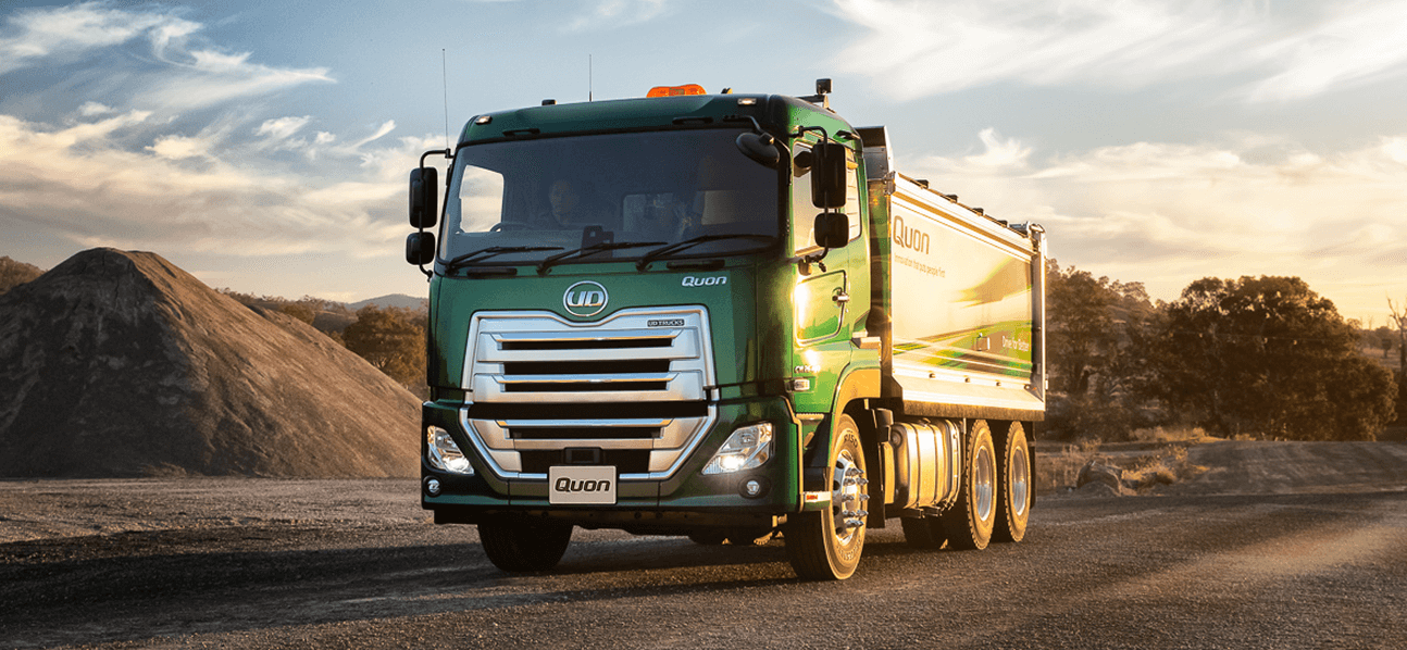 Green dump truck on a dirt road, with gravel piles and trees in the background.